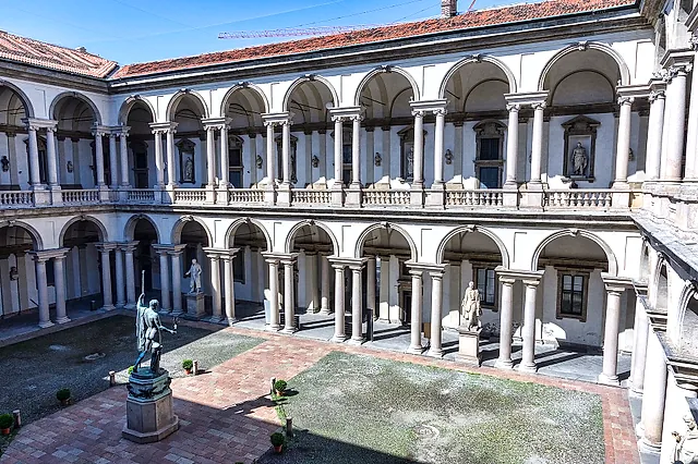 The courtyard of Brera Palace with two levels of columned arcades in pale stone, a bronze statue on a pedestal in the foreground and a brick-paved floor.