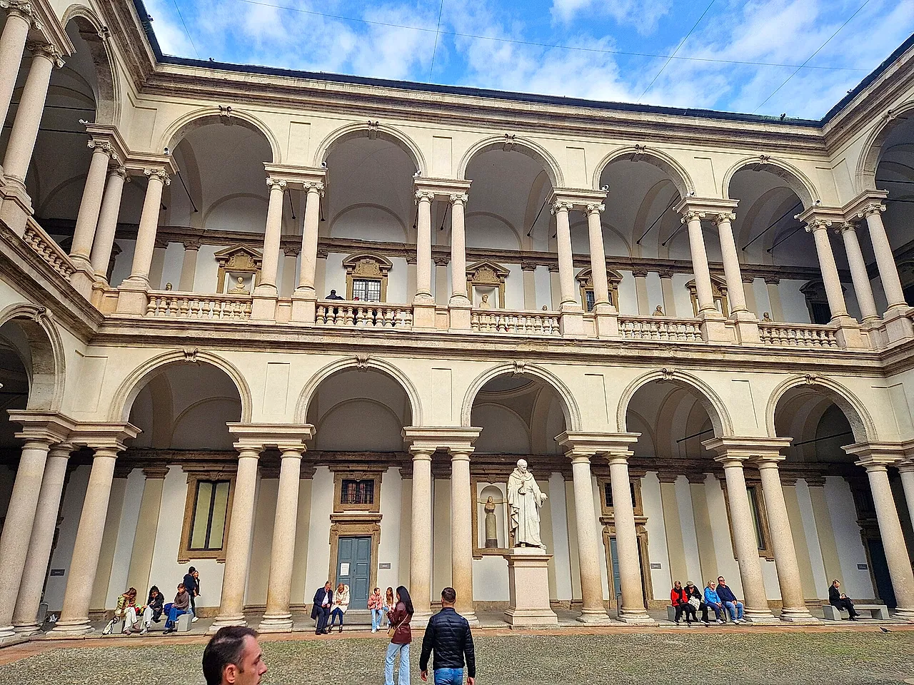 The courtyard of Brera Palace with white arcades on two levels and a white statue on a pedestal at the centre; visitors stand in the foreground.