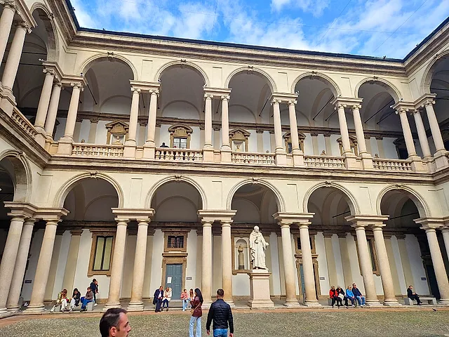 The courtyard of Brera Palace with white arcades on two levels and a white statue on a pedestal at the centre; visitors stand in the foreground.