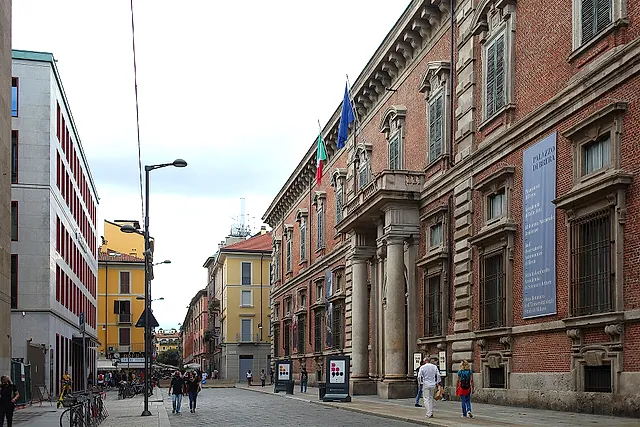 The red-brick facade of Brera Palace along a city street, with a stone portal, flags on the upper floor and a large banner on the wall; pedestrians walk the sidewalk.