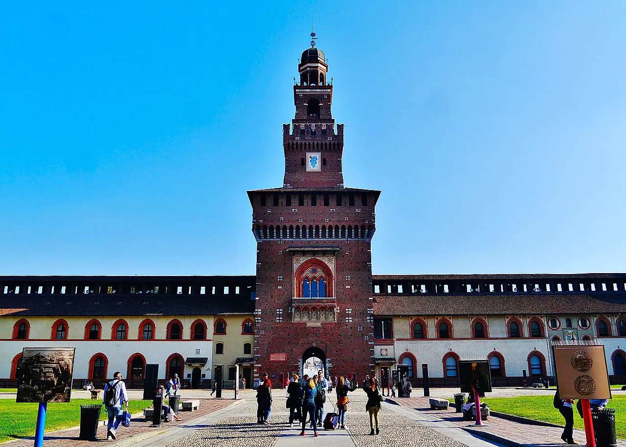The central gate tower of Sforza Castle seen from the forecourt, with its arched entrance and cream buildings with red-arched windows on either side, and visitors walking across the paved courtyard.