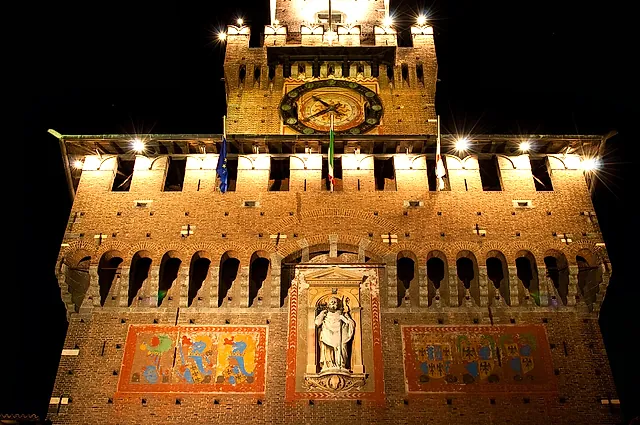 A brick tower of Sforza Castle lit up at night, with a clock in its upper part and a stone statue in a central niche between two decorated panels, against a dark sky.