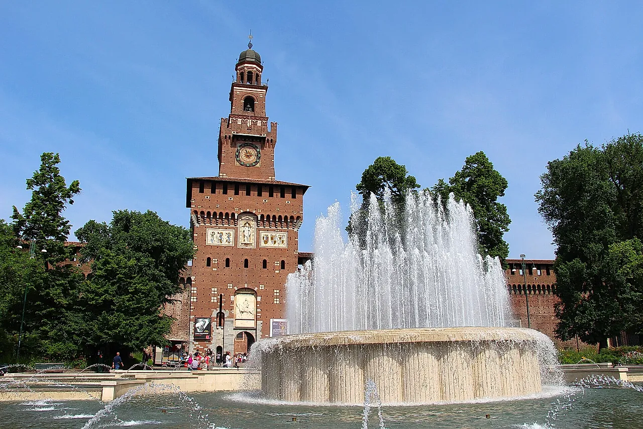 The clock tower of Sforza Castle seen across a fountain, its jets of water rising in front of the red-brick walls under a clear blue sky.