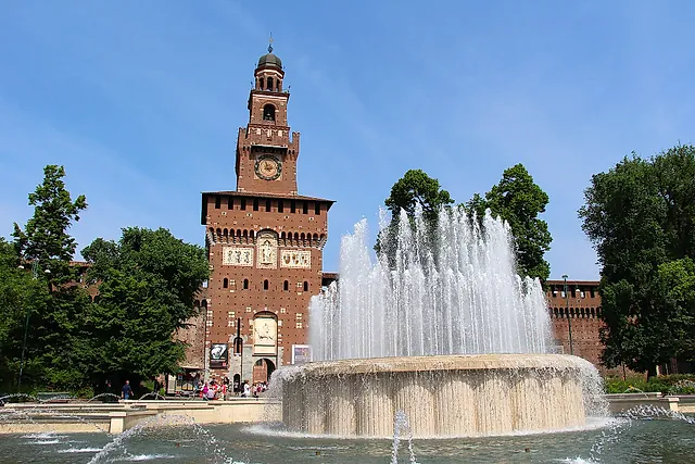 The clock tower of Sforza Castle seen across a fountain, its jets of water rising in front of the red-brick walls under a clear blue sky.