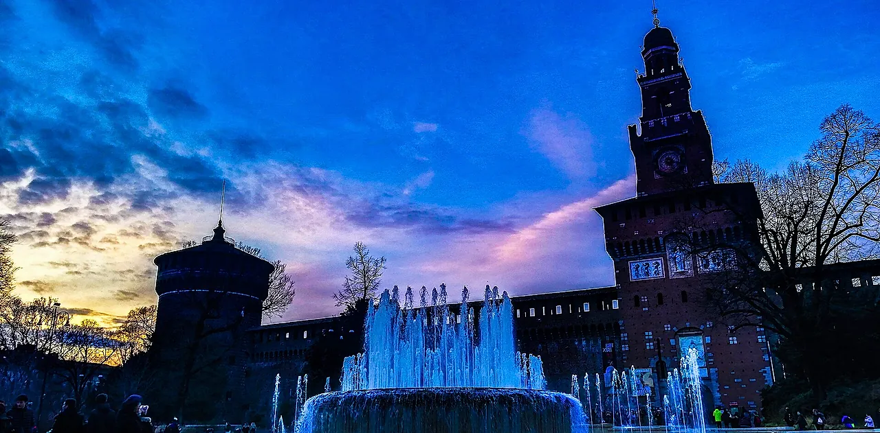 Sforza Castle at dusk, its brick tower and walls silhouetted against a blue and pink sky, with a lit fountain throwing water up in the foreground.