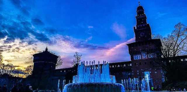 Sforza Castle at dusk, its brick tower and walls silhouetted against a blue and pink sky, with a lit fountain throwing water up in the foreground.