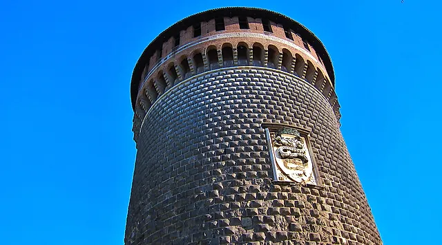 The round tower of Sforza Castle, built of dark stone and brick, seen from below under a clear blue sky, with a carved stone coat of arms set in its wall.