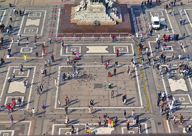 Aerial view of the Piazza del Duomo from the cathedral roof: the light geometric paving, a crowd of visitors and pigeons, and a stone monument at the top of the frame.