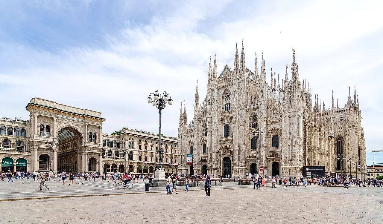 The Galleria Vittorio Emanuele II arch on the left and the cathedral facade on the right across a wide piazza, with pedestrians walking in the square.