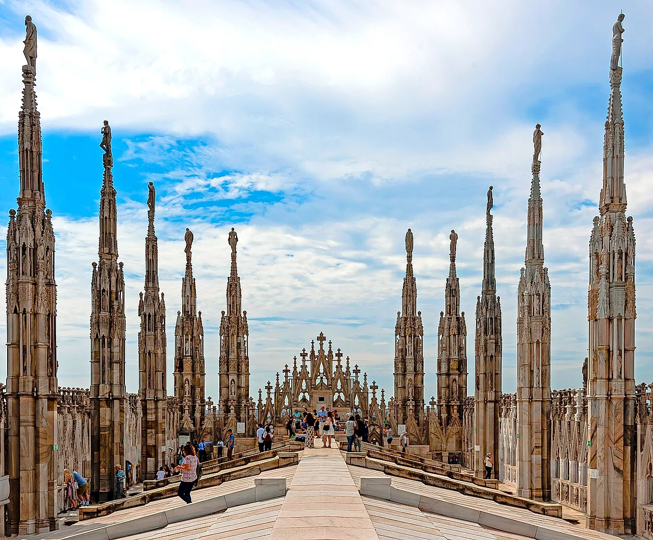 The cathedral roof terraces, lined with rows of marble spires topped by statues, with a central walkway leading toward the facade under a cloudy sky and a few visitors on the path.