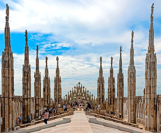 The cathedral roof terraces, lined with rows of marble spires topped by statues, with a central walkway leading toward the facade under a cloudy sky and a few visitors on the path.