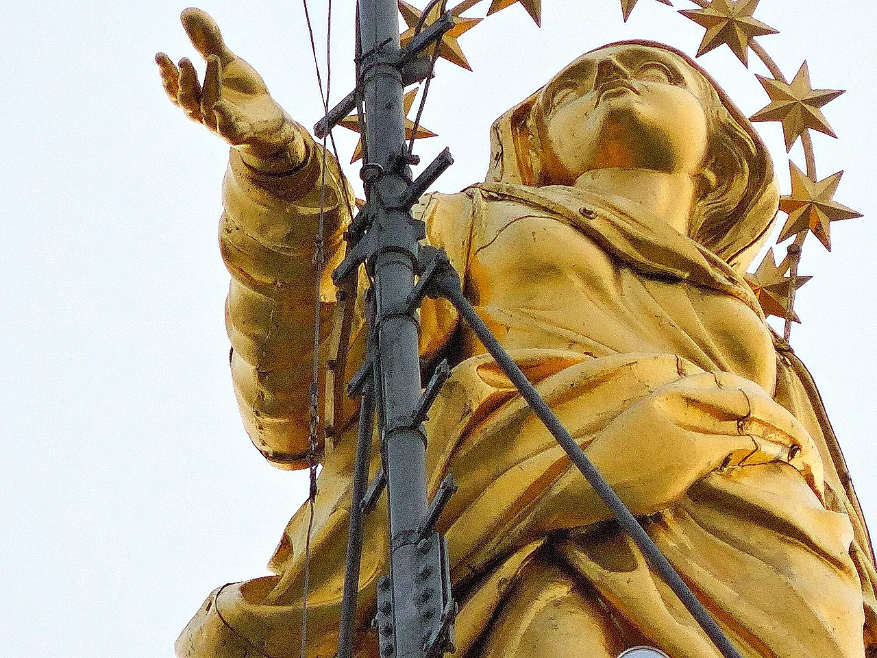 The gilded Madonnina statue on top of the cathedral spire, seen from below against a pale sky, with its outstretched arm and star halo, and a metal lightning rod with cables in front of it.