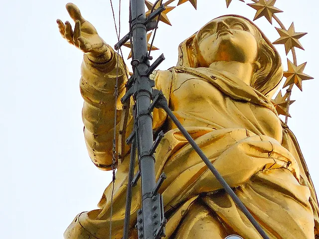 The gilded Madonnina statue on top of the cathedral spire, seen from below against a pale sky, with its outstretched arm and star halo, and a metal lightning rod with cables in front of it.