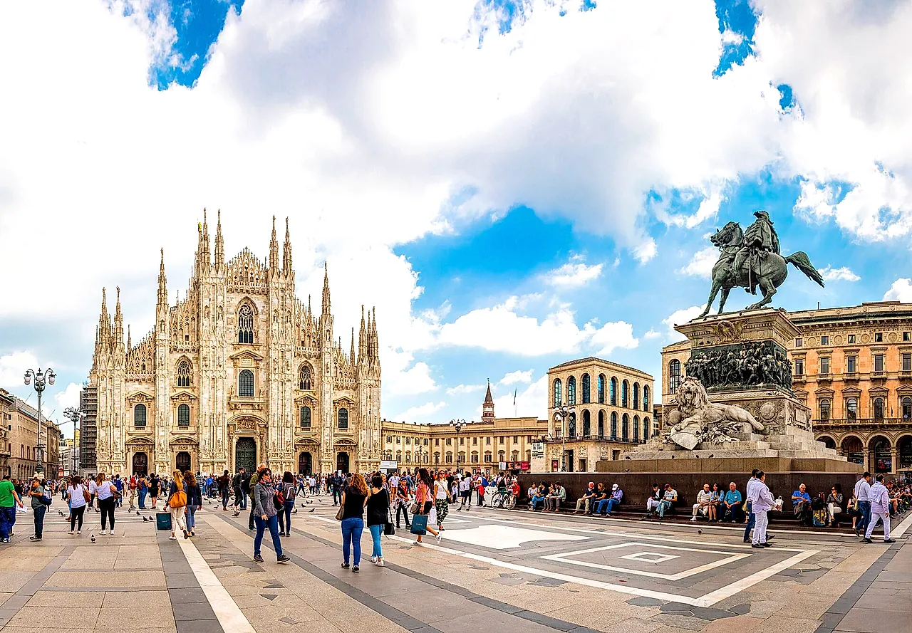 Piazza del Duomo with the cathedral on the left and the equestrian statue of Victor Emmanuel II on the right, a wide sky with clouds, and passers-by in the foreground.