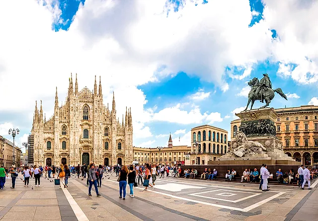 Piazza del Duomo with the cathedral on the left and the equestrian statue of Victor Emmanuel II on the right, a wide sky with clouds, and passers-by in the foreground.
