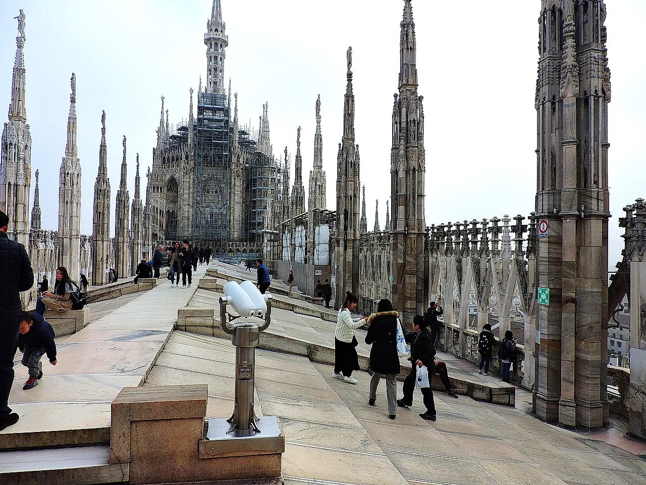 A rooftop walkway of the Milan Cathedral with statue-topped spires and scaffolding on part of the cathedral in the distance, with visitors on the terrace and a coin-operated viewer in the foreground.