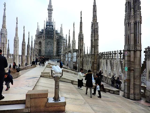A rooftop walkway of the Milan Cathedral with statue-topped spires and scaffolding on part of the cathedral in the distance, with visitors on the terrace and a coin-operated viewer in the foreground.