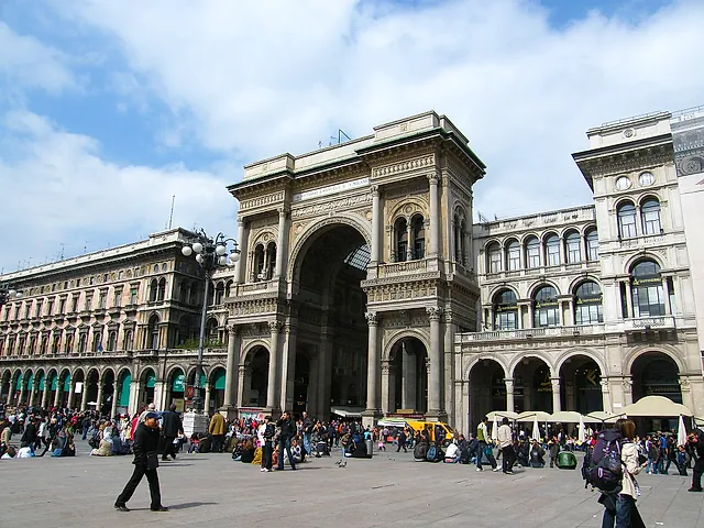 The monumental triumphal arch at the entrance of the Galleria Vittorio Emanuele II from Piazza del Duomo, with pedestrians and a few people in the foreground under a cloudy sky.