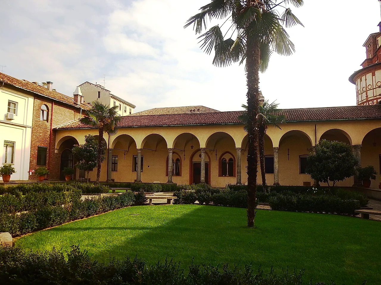 The cloister of Santa Maria delle Grazie: a cream-coloured arcade around a lawn, a tall palm tree in the middle and clipped hedges along the borders.
