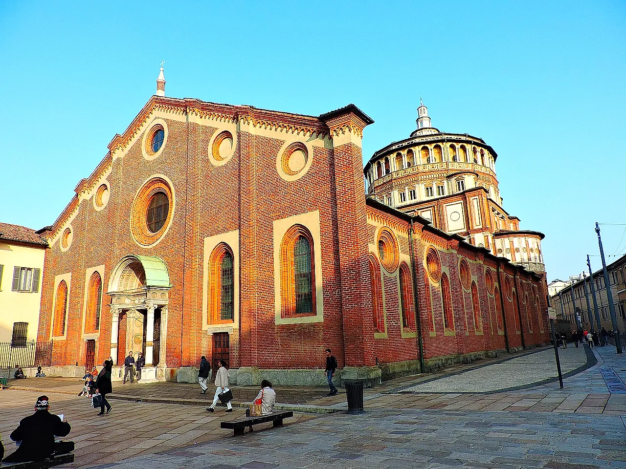 The brick facade of Santa Maria delle Grazie on a sunny square, with a pale stone portal, arched windows and the round drum of the dome visible at right.