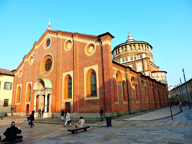 The brick facade of Santa Maria delle Grazie on a sunny square, with a pale stone portal, arched windows and the round drum of the dome visible at right.