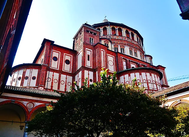 The red-brick apse of Santa Maria delle Grazie seen from the north, with a domed drum above rows of round windows, framed by a leafy tree in the foreground.