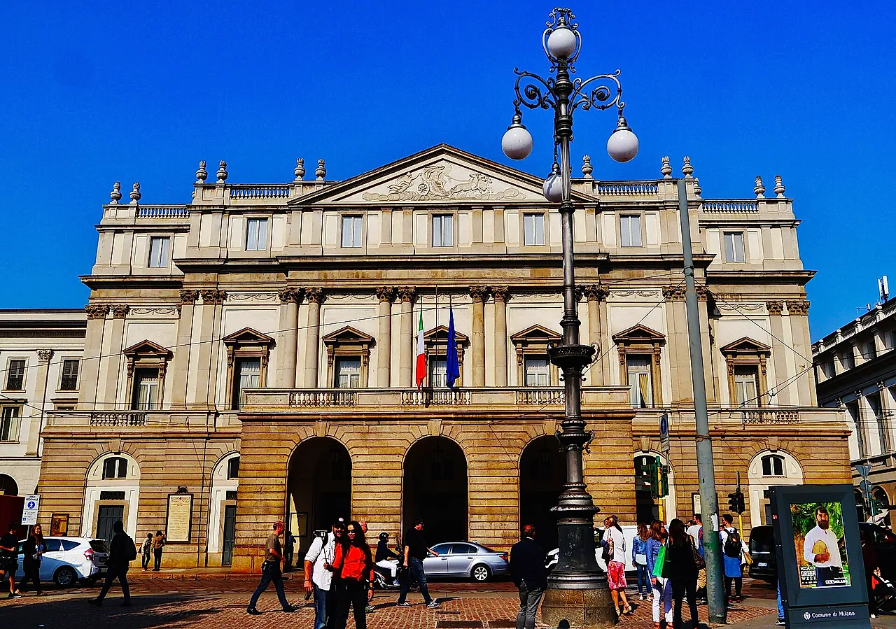 The neoclassical facade of the La Scala opera house, with columns, a triangular pediment and arched entrances at street level, seen from the square with passers-by.