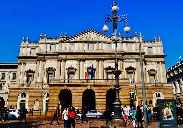 The neoclassical facade of the La Scala opera house, with columns, a triangular pediment and arched entrances at street level, seen from the square with passers-by.