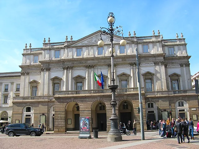La Scala seen from the square, with its arched entrances and a wrought-iron street lamp in the centre, and people walking in the foreground on the right.