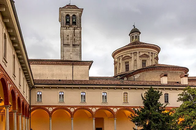 The cloister of the science museum: a cream-coloured building with an arcade lit by warm light on the left, and a bell tower and a domed drum rising behind the roofs under a cloudy sky.