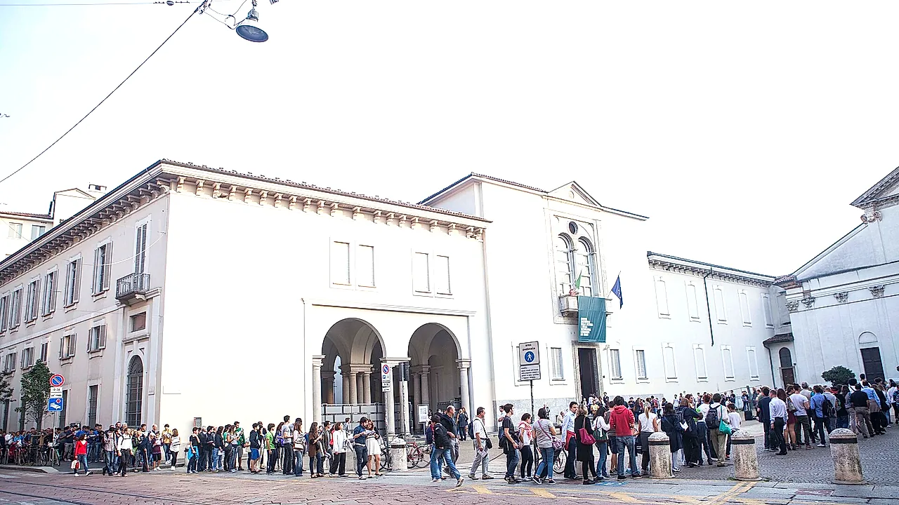 The white facade of the science museum entrance, with a loggia of arches at ground level and a long line of visitors waiting outside under an overcast sky.