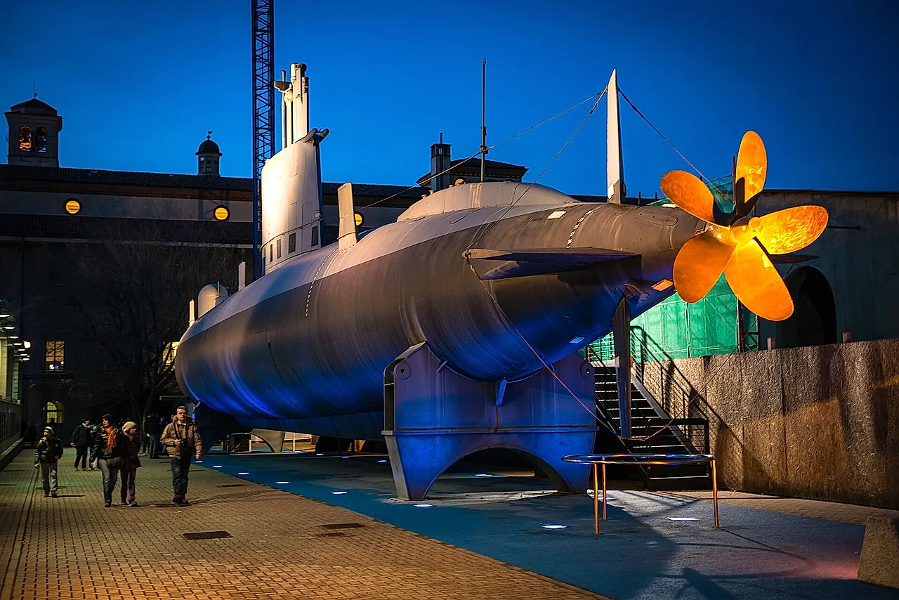 The submarine Enrico Toti displayed outdoors at dusk, its grey hull lit by blue floodlights and its golden propeller glowing at right, with a few visitors at left.