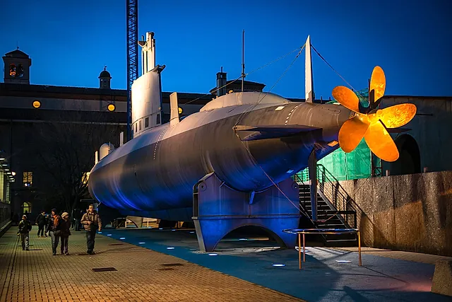 The submarine Enrico Toti displayed outdoors at dusk, its grey hull lit by blue floodlights and its golden propeller glowing at right, with a few visitors at left.