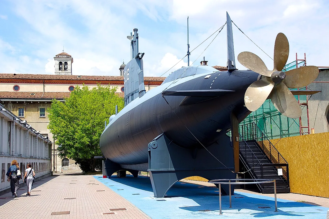 The submarine Enrico Toti in daylight, seen from the side with its grey hull and bronze-coloured propeller at right, on a paved courtyard with two visitors at left.