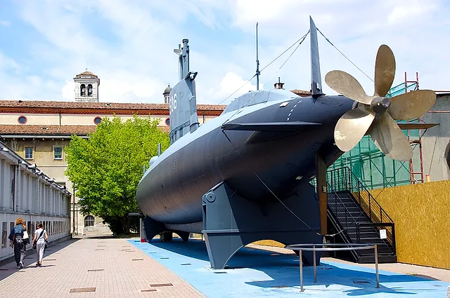 The submarine Enrico Toti in daylight, seen from the side with its grey hull and bronze-coloured propeller at right, on a paved courtyard with two visitors at left.